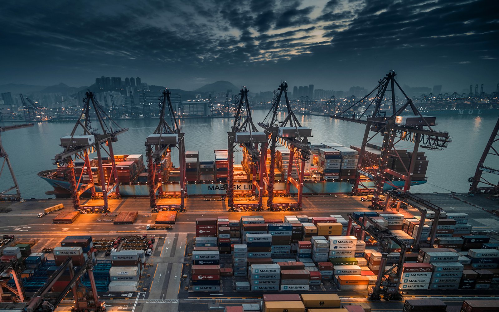 Gantry cranes loading a container ship at a port at dusk, stacked containers filling the yard